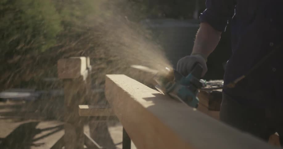 A man shaves a wood beam outdoors