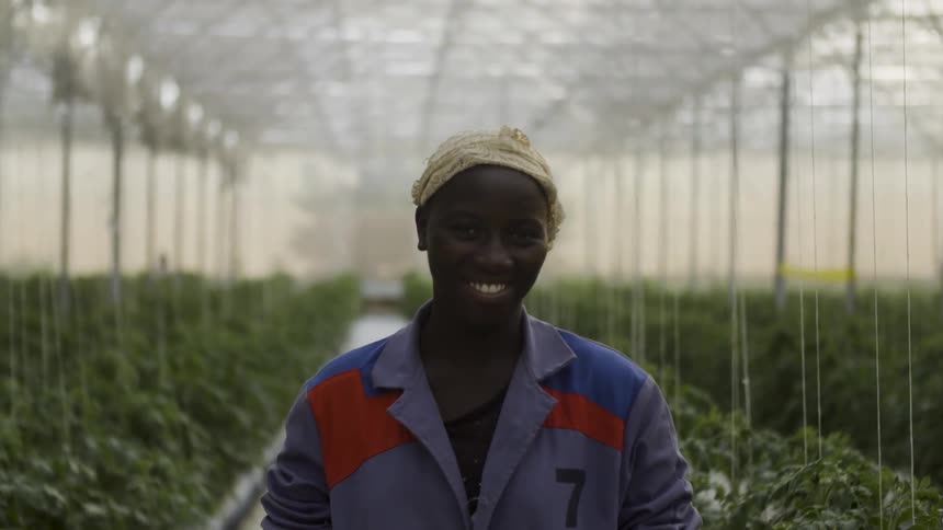 A woman stands smiling among a row of plants inside a greenhouse