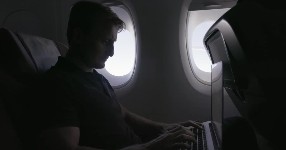 A man is sitting in a dark airplane using his laptop