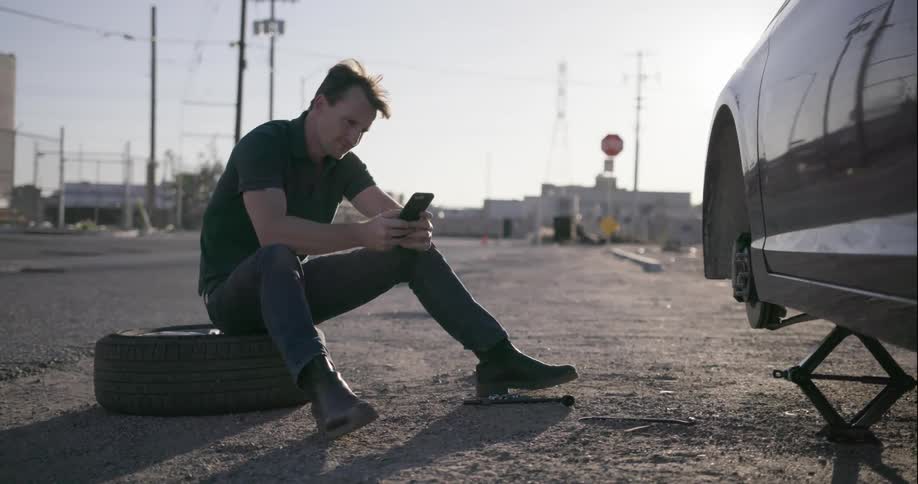 A man sits on a spare tire and types on a smartphone next to a car with a flat