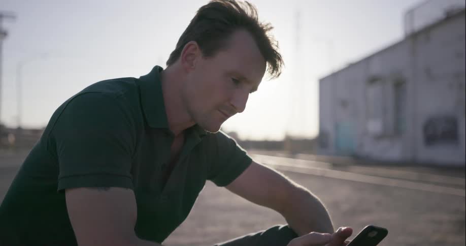 A man types on a smartphone while sitting on the ground outside