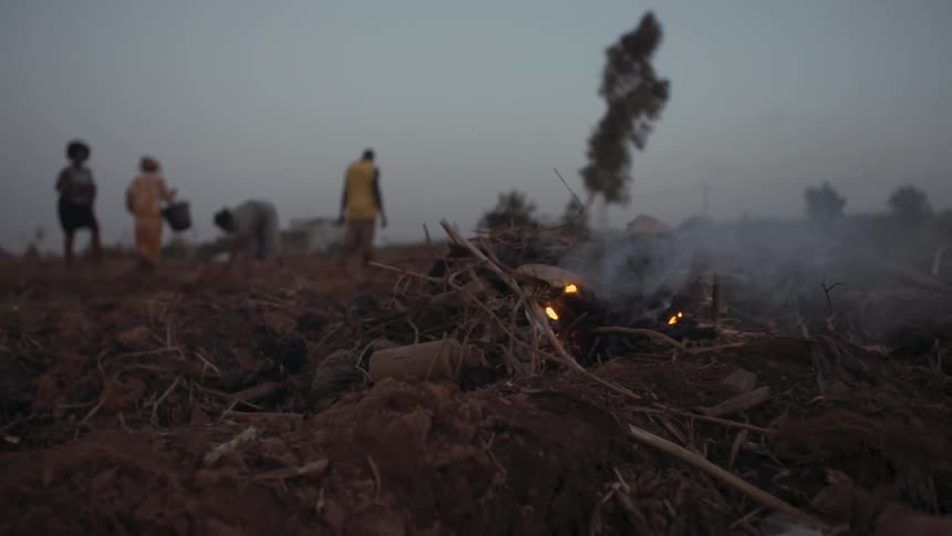 A fire burns in a rural field with workers in the distance