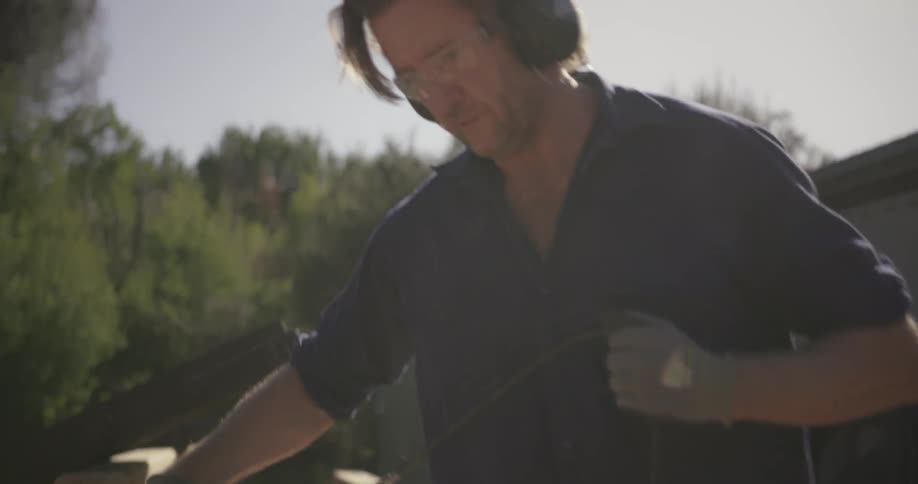 A man shaves a wood beam outdoors