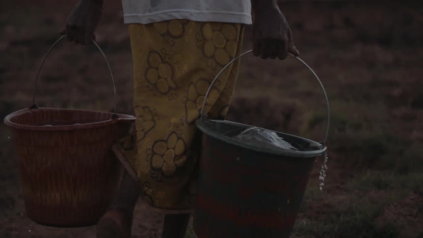 Woman carrying two buckets full of water outside through dirt