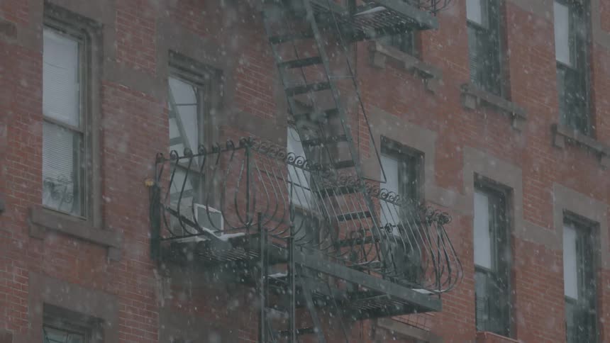 Snow is falling around a brick building that has small balconies and ladders on each floor