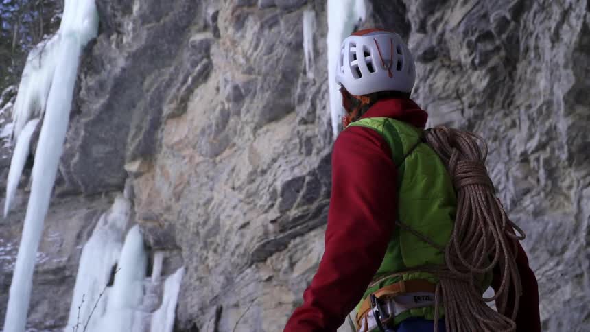 Man wearing ice climbing gear is standing and staring towards rock wall