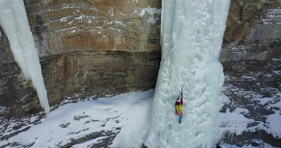 Man wearing helmet and ropes using ice axes to climb up frozen waterfall