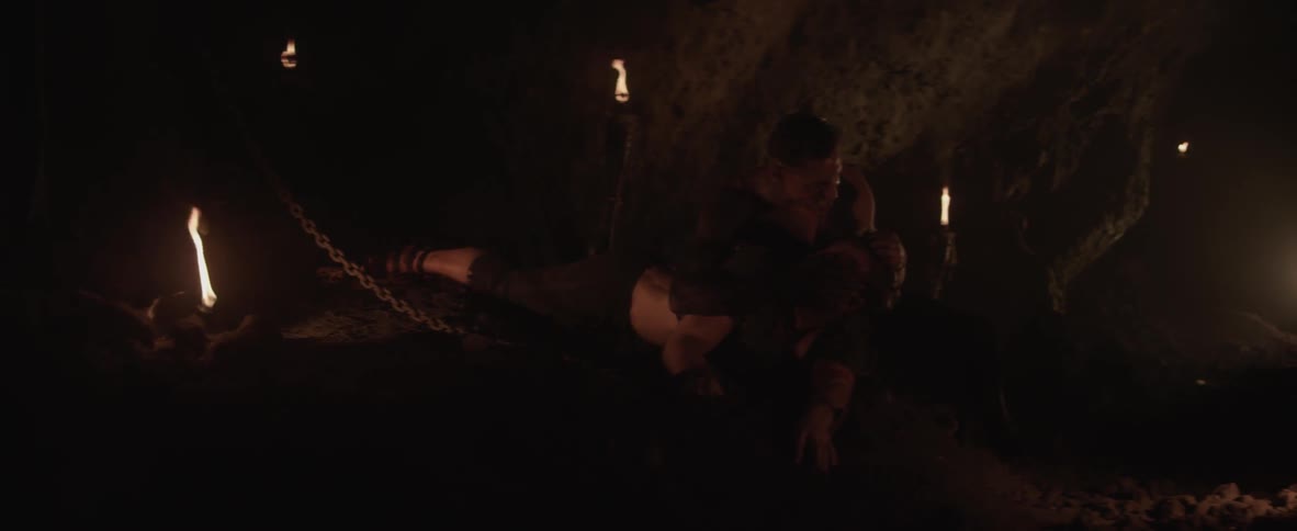 Man holds another man's head in his arms in a cave lit by fire