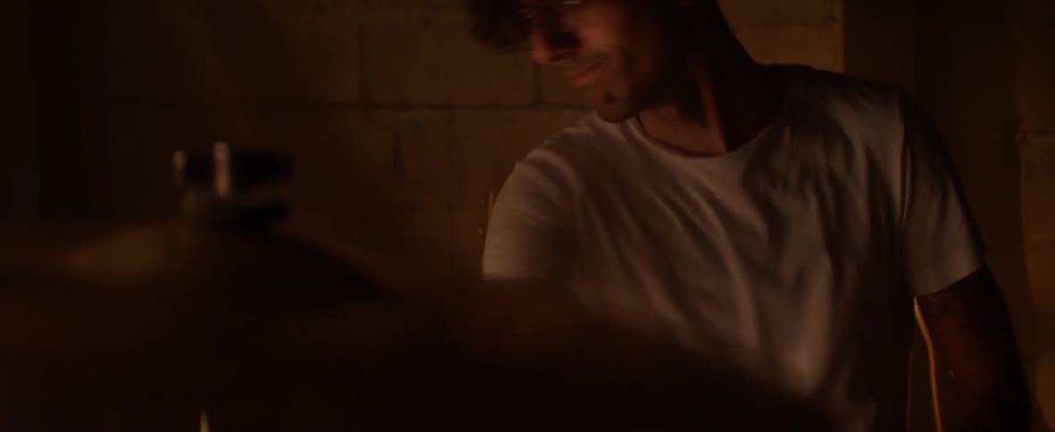 Man in white shirt plays drums in a dimly lit room