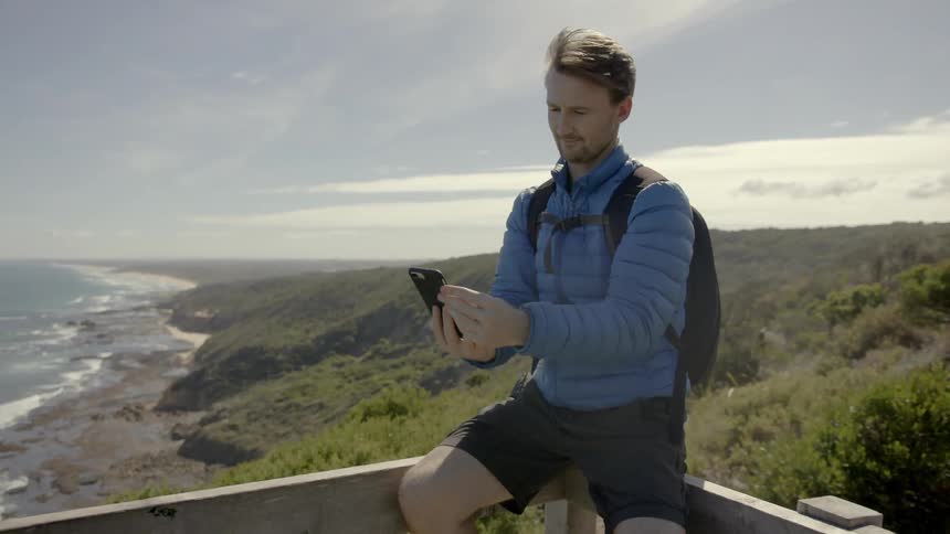 Man sitting high on a rail and taking pictures with his cell phone next to the sea on a cloudy day