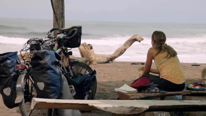 A woman eats on a bench on a beach while looking out at the ocean