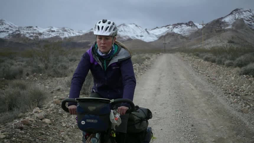 A woman bicycles on a rural dirt road with snowy mountains in the distance