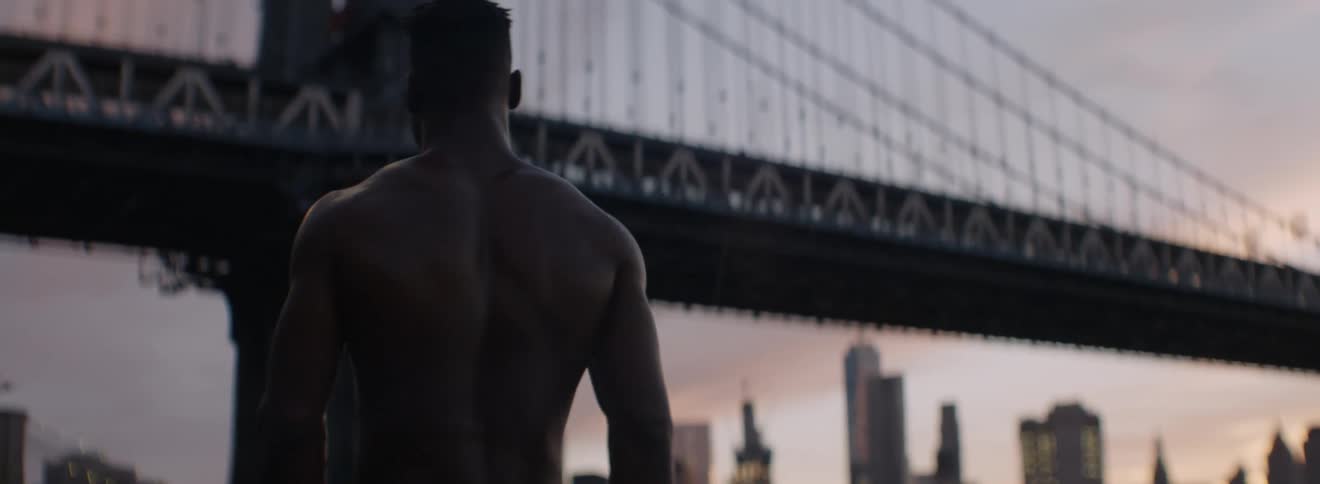 A shirtless man walks next to a river flowing through a city at sunset
