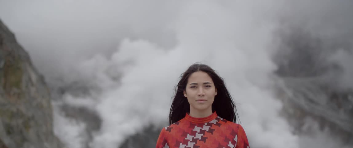 A woman is standing and smiling while a volcano behind her vents steam