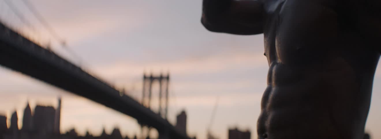 A shirtless man drinks from a water bottle in a city at sunset