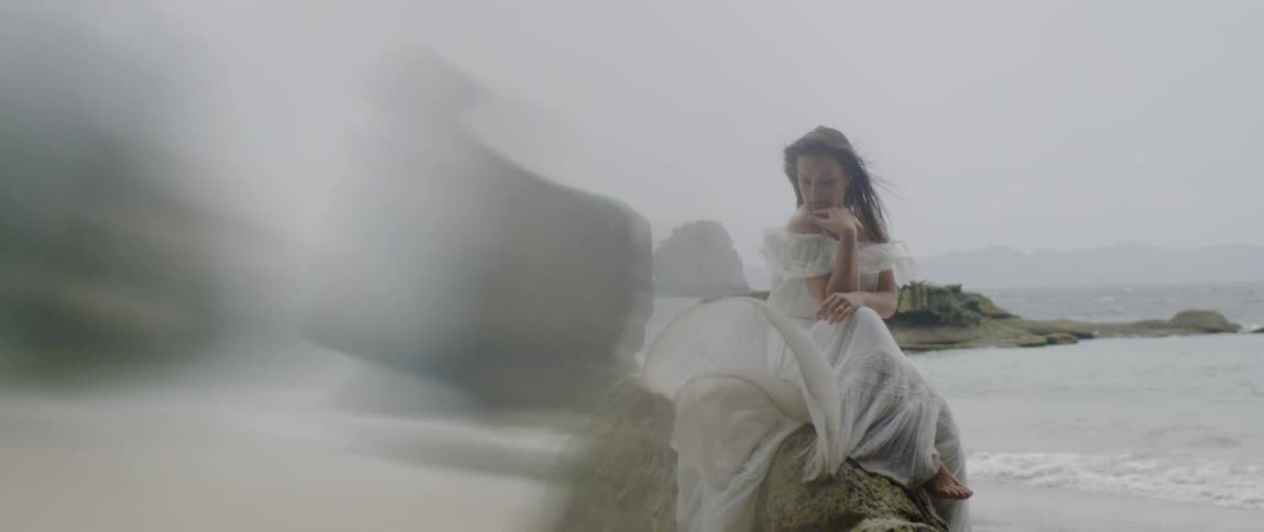 A woman in a white dress sits on a rock on the beach