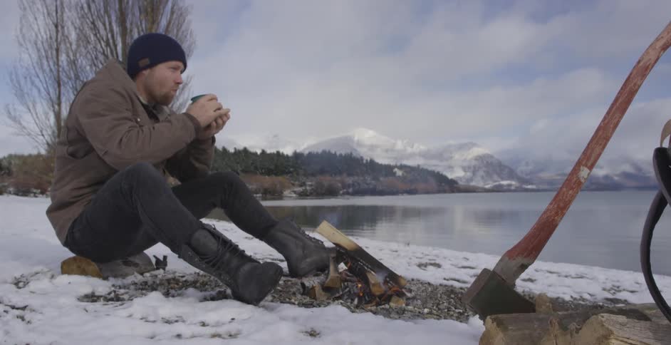 Man sitting lakeside on a snowy day drinking from a cup in front of his campfire
