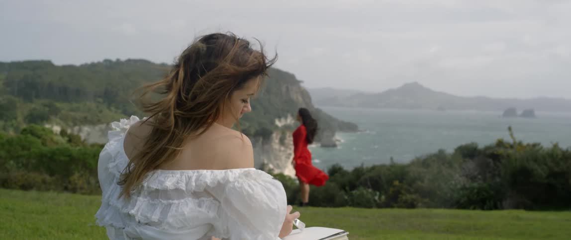 A woman in white writes in a book as another in red looks over the water
