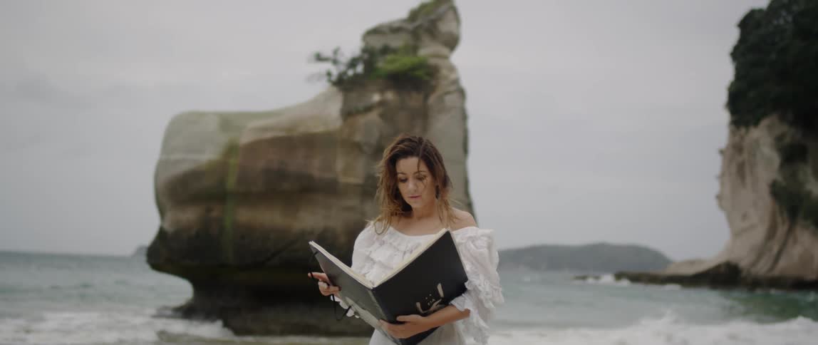 Woman holding a large black binder stands in front of the ocean