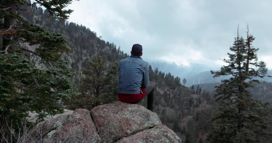 Man climbs up on a rock and sits looking at the mountains