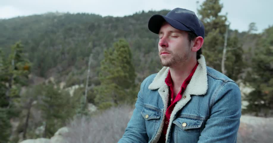 Man wearing a hat and a denim jacket sits on a mountain breathing deeply