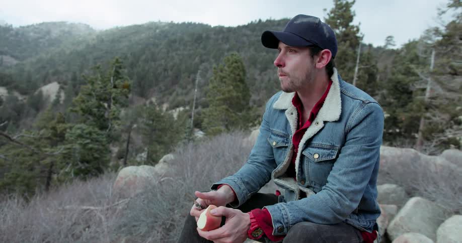 A man cuts an apple with a knife and eats it while sitting in the mountains