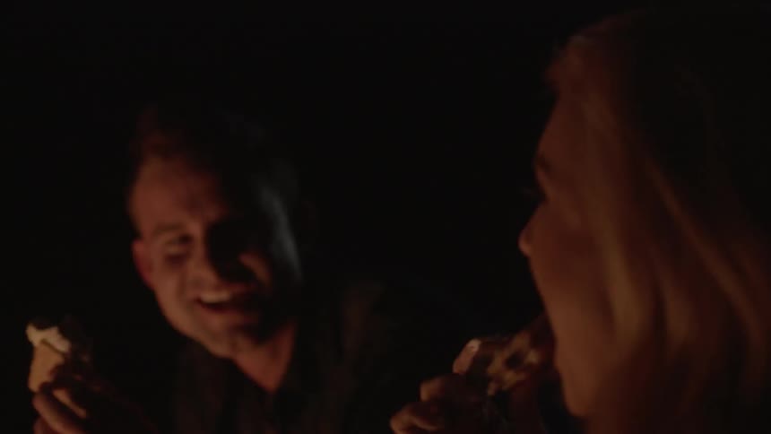 A man and woman kiss as they eat s'mores next to a campfire