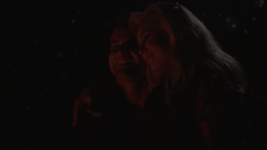 A man and woman hug in front of a fire at night
