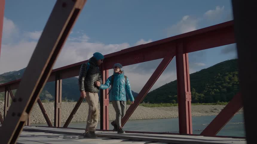 A man and woman hold hands while crossing a river bridge in the mountains