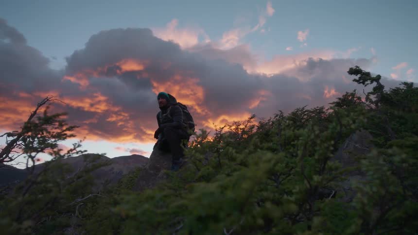 A man stands on a very windy hill at sunset