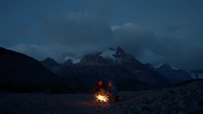 Two people sit on the beach in front of a campfire