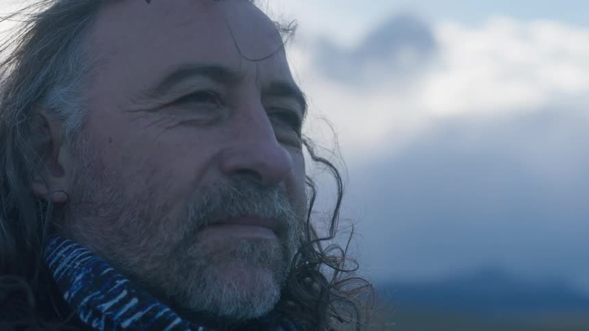 A man stares off into the distance with cloudy mountains behind him