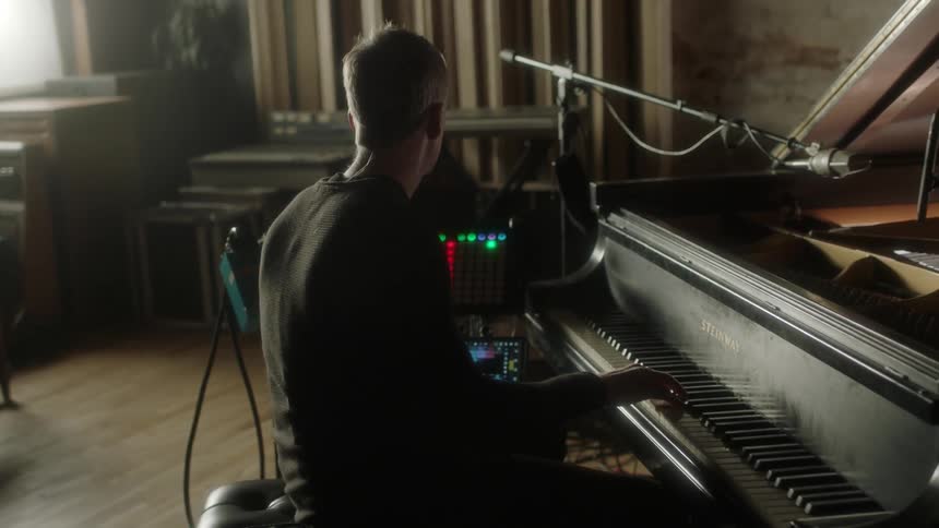 A musician sits at a computer and midi controllers