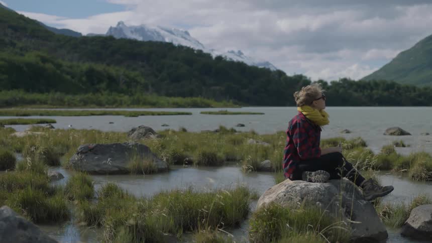 A woman sits on a rock by the water on a windy day