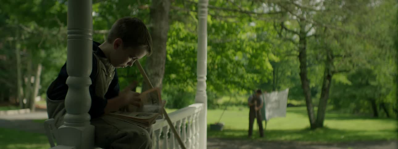 A boy sits on a porch rail while a man works in the yard