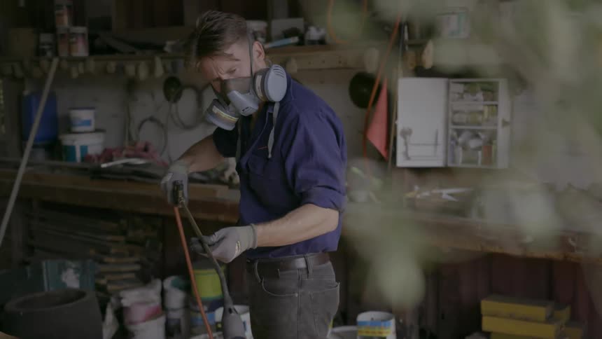 Man wearing a gas mask uses a sprayer to spray some wood