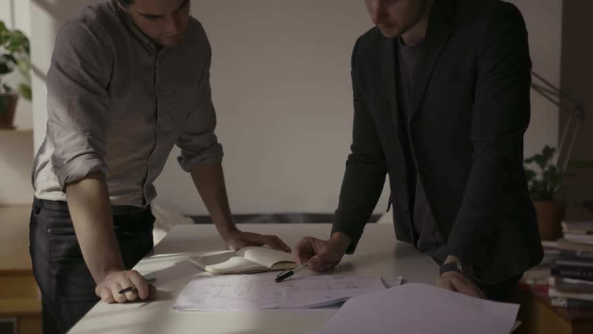 Two men stand at a desk pointing at sheets of paper