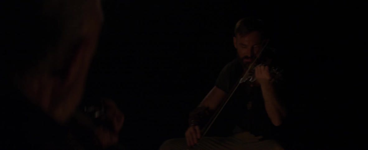 A man is sitting by a camp playing the violin