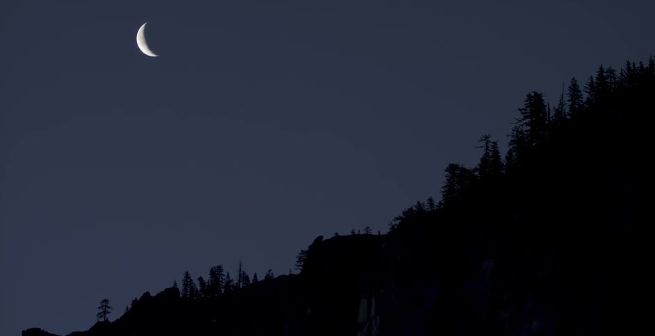 A sliver of the moon is visible next to the dark woods on a hill
