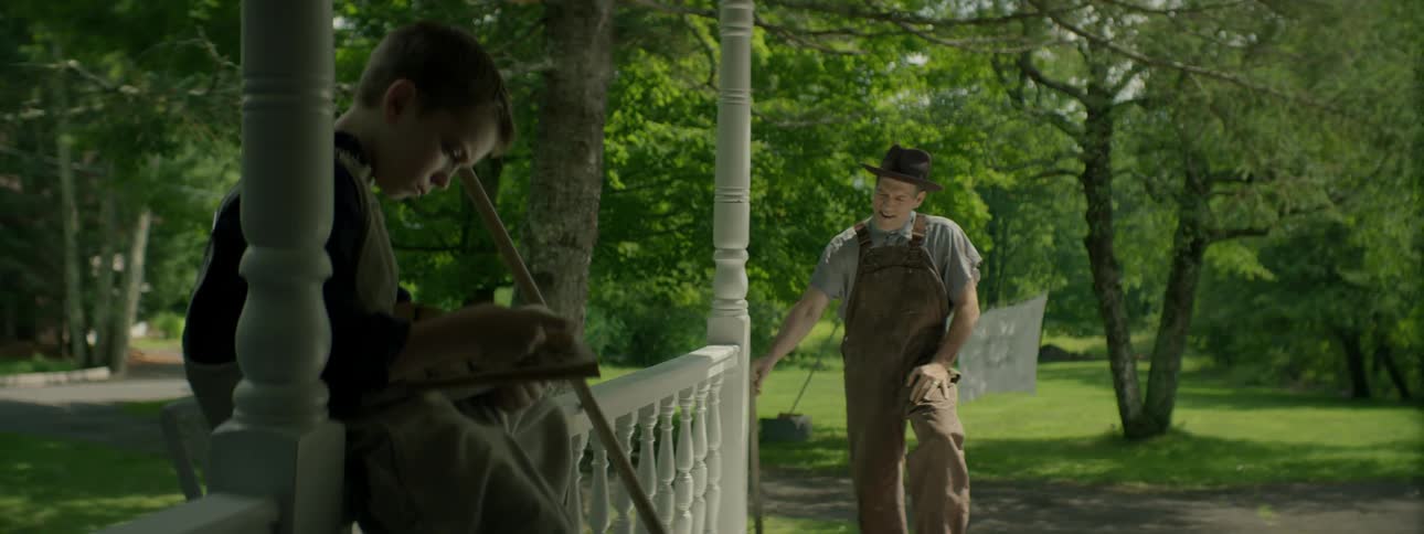 A farmer approaches his son who is reading a book on the porch