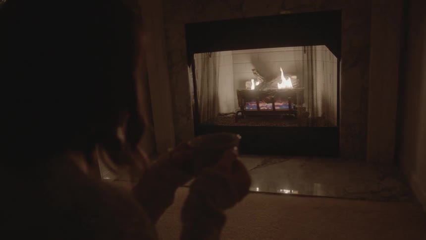 A woman holding a coffee cup gazes into the fire in a fireplace