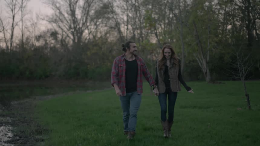 A man and woman hold hands and walk on the grass near a pond