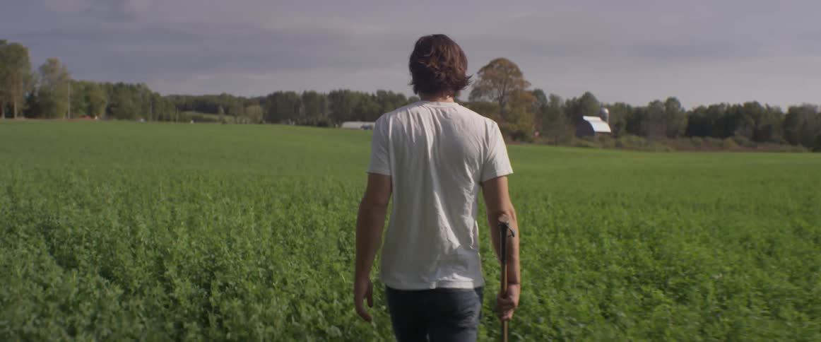 Man holding bat walking through tall green grass in field on cloudy day