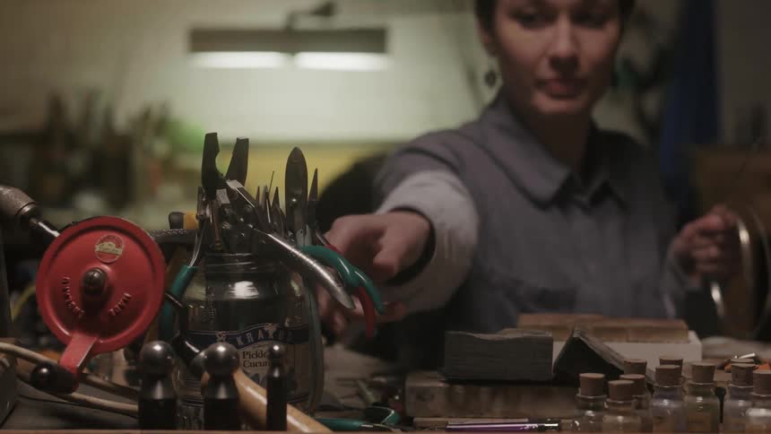 A woman sits on a workbench lifting up a pair of pliers