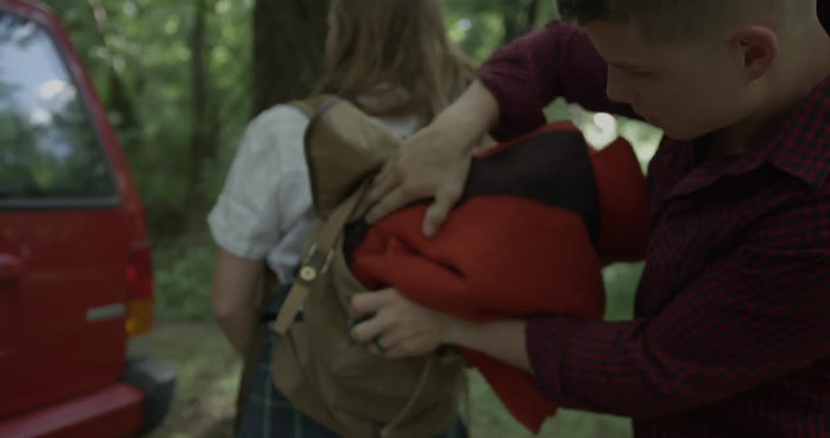 A man is putting a blanket into a backpack a woman is carrying