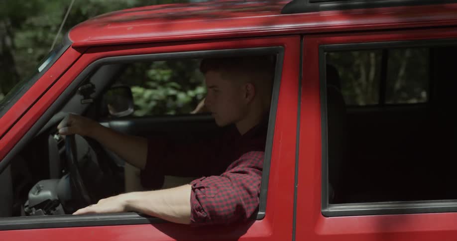 A man drives a red car past trees