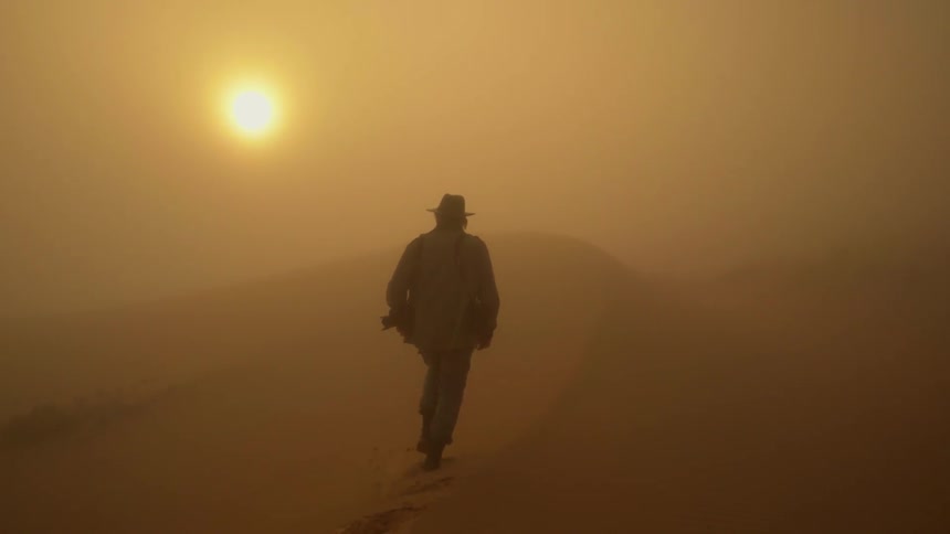 A man is walking along the dunes in the desert with the sun blazing down in front of him