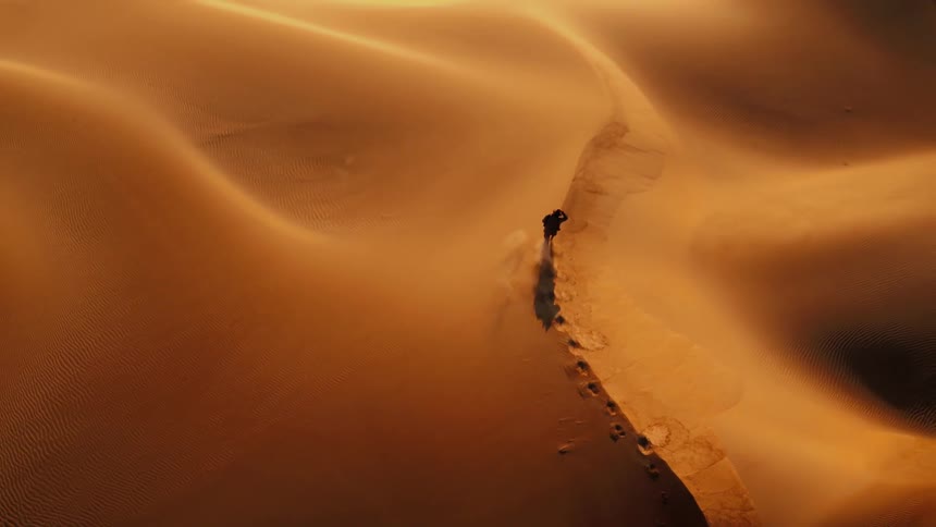 A man is walking along sand dunes being filmed from a drone
