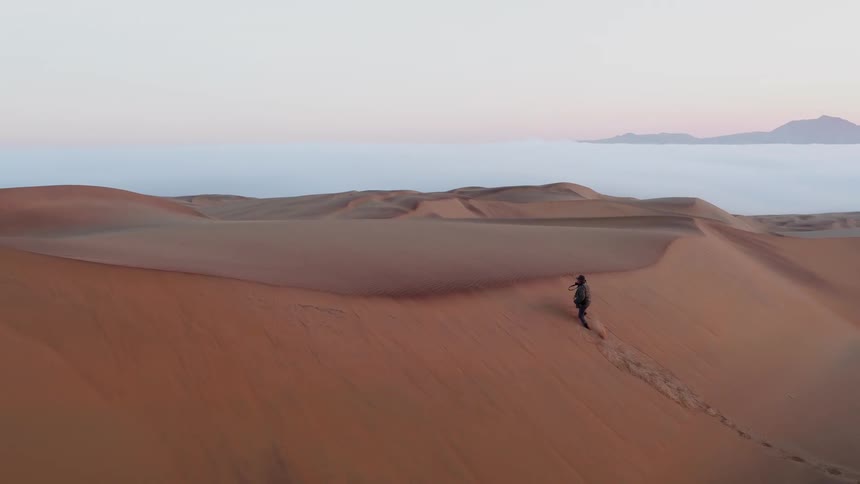 A photographer climbs up a sand dune in a desert at sunset
