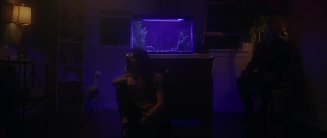 A man sits on the floor and leans against a fish tank in a dark room